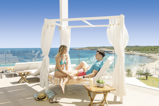 Mann und Frau sitzen auf einem Daybed auf einer Sonnenterasse eines Hotels, im Hintergrund das Meer